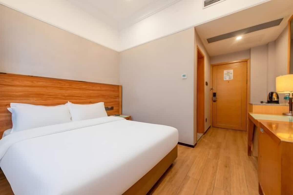 A hotel room with a wooden headboard, a bed with white linens, a wooden desk with a lamp, and a door leading to another room.