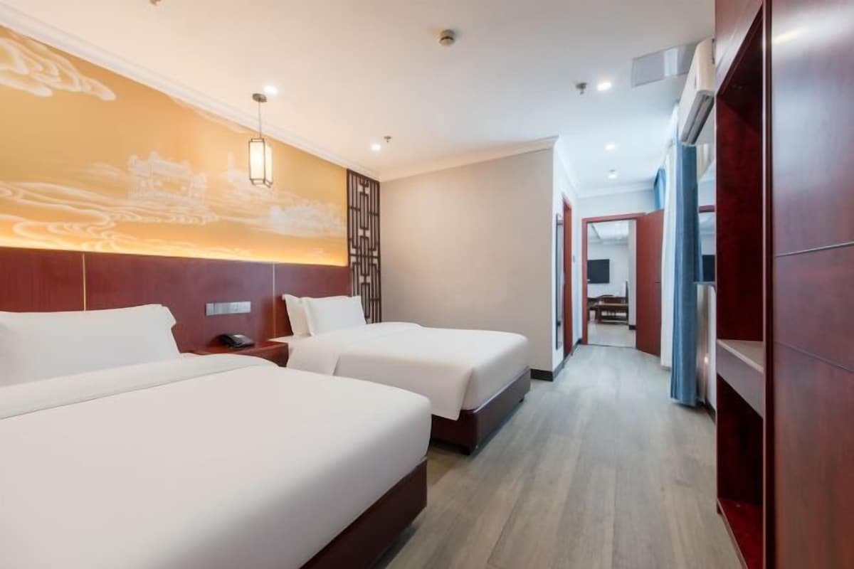 A hotel room with two beds, a wooden headboard, a wall-mounted light fixture, and a view of the hallway.