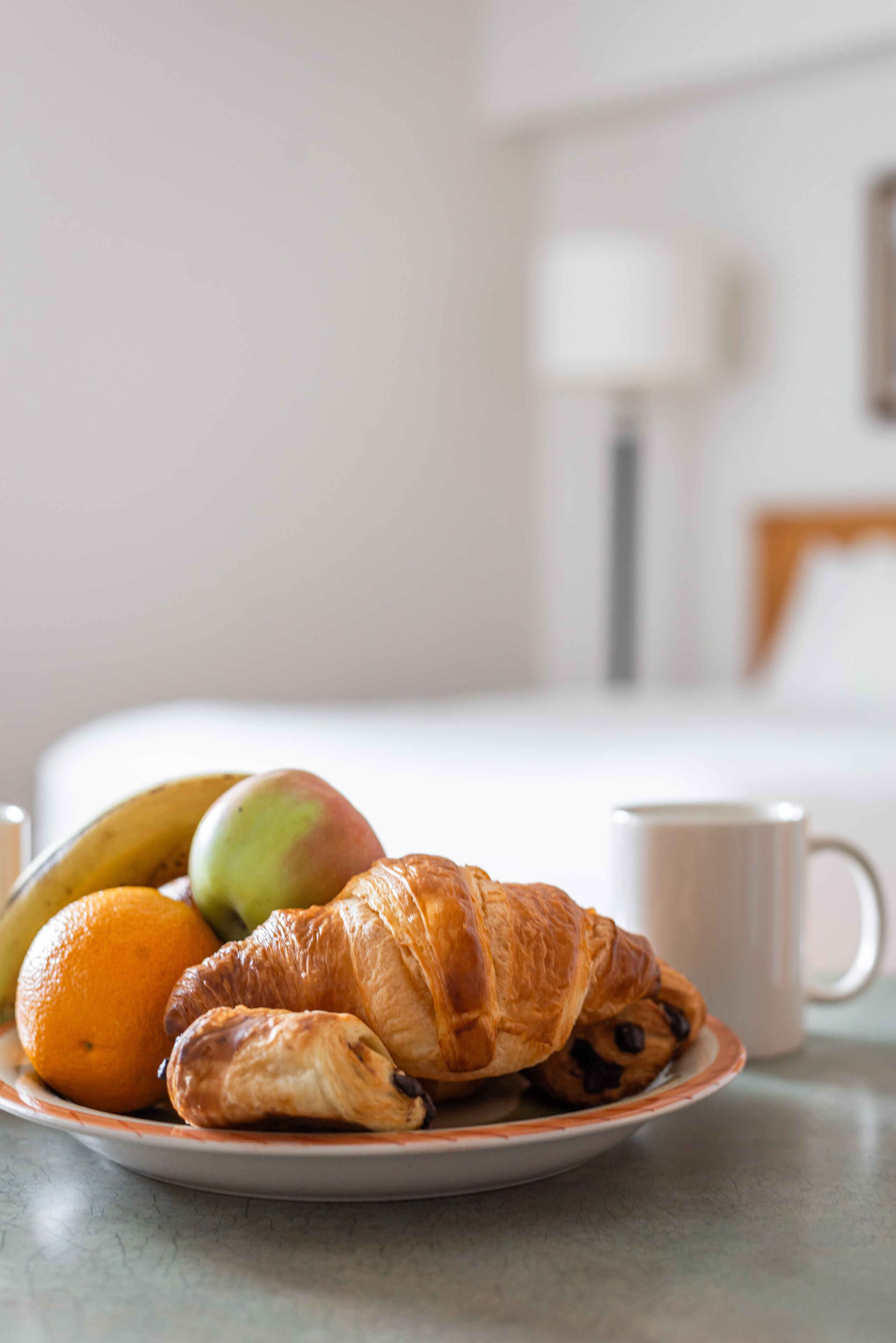 A plate with a croissant, an orange, and a green apple on a table in a hotel room.