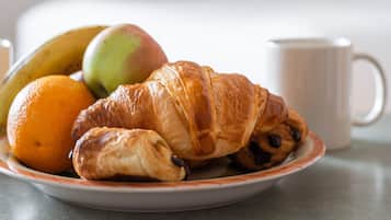 A plate with a croissant, an orange, and a green apple on a table in a hotel room.