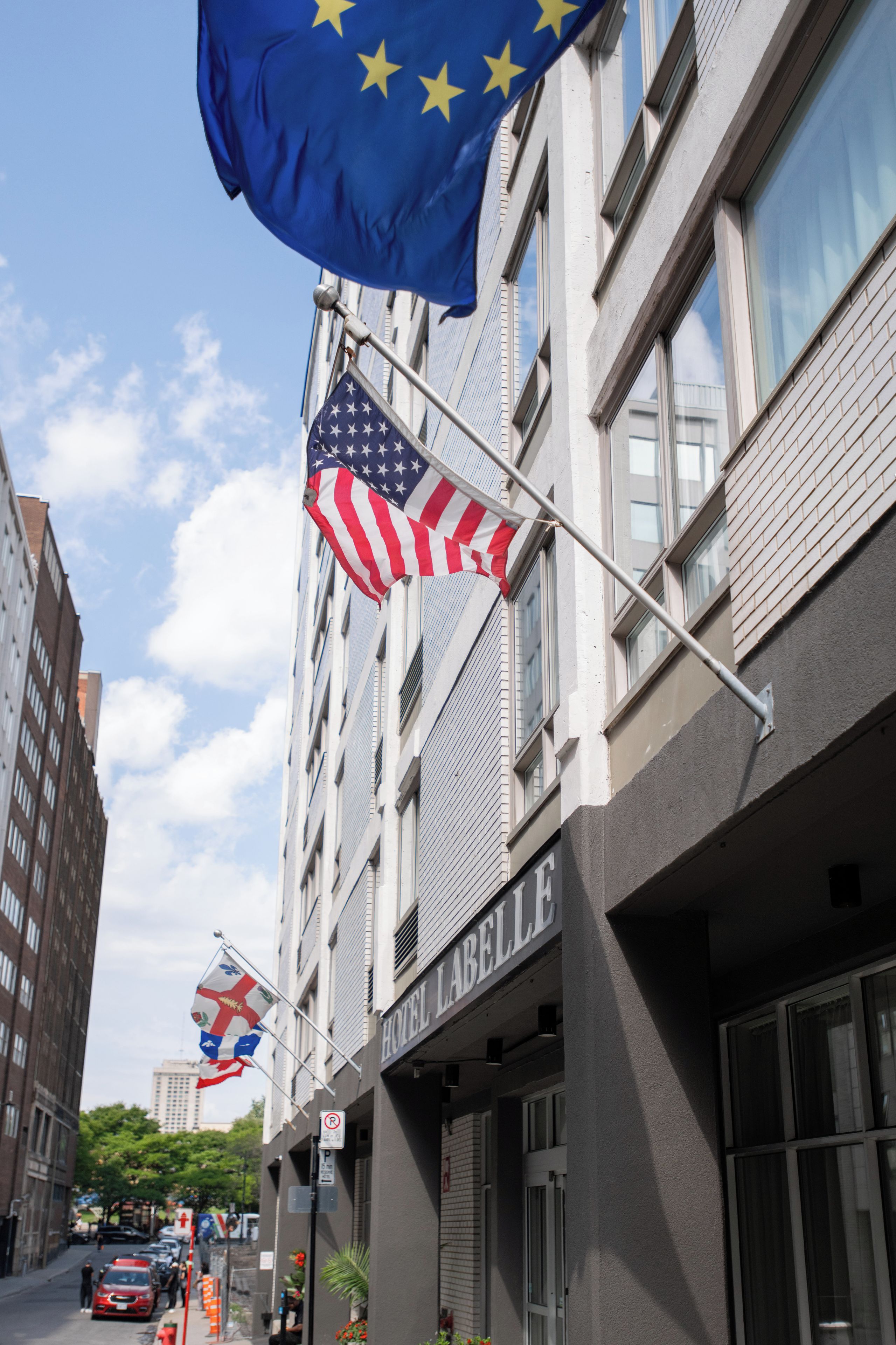 Un bâtiment sur lequel on peut lire l’inscription « HÔTEL L’ABELLE » et qui est orné de plusieurs drapeaux, dont le drapeau américain et le drapeau de l’Union européenne.