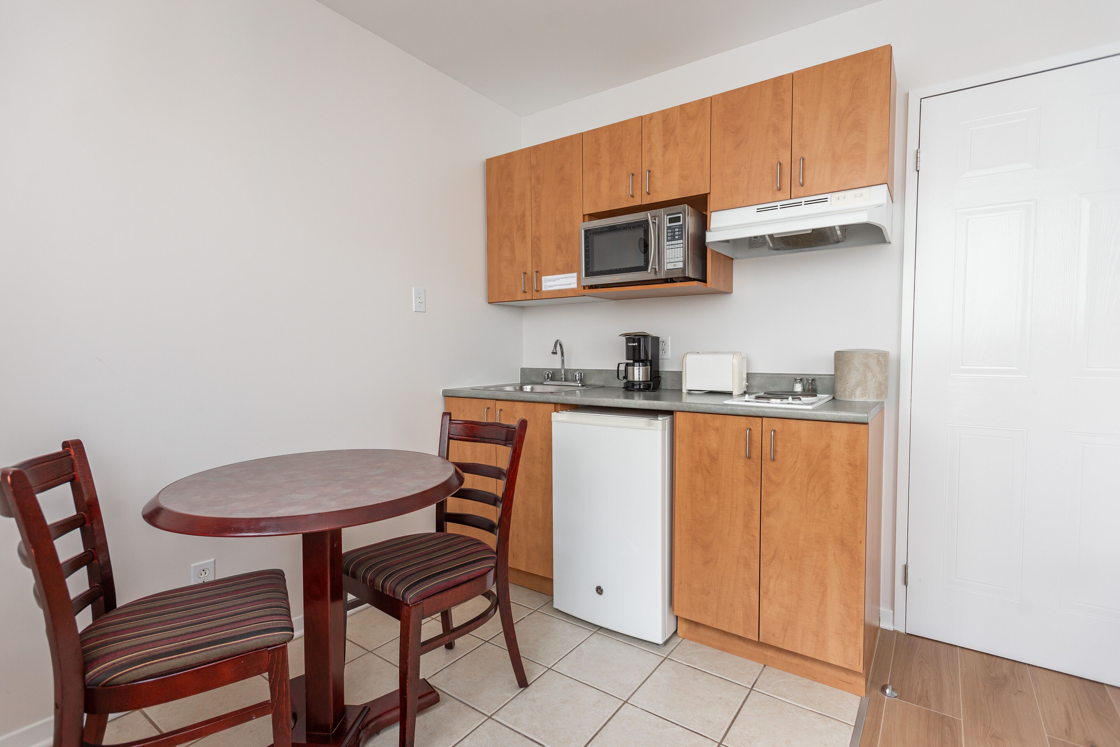 A compact kitchen with wooden cabinets, a microwave, a coffee maker, and a small refrigerator. There is a round table with two chairs.