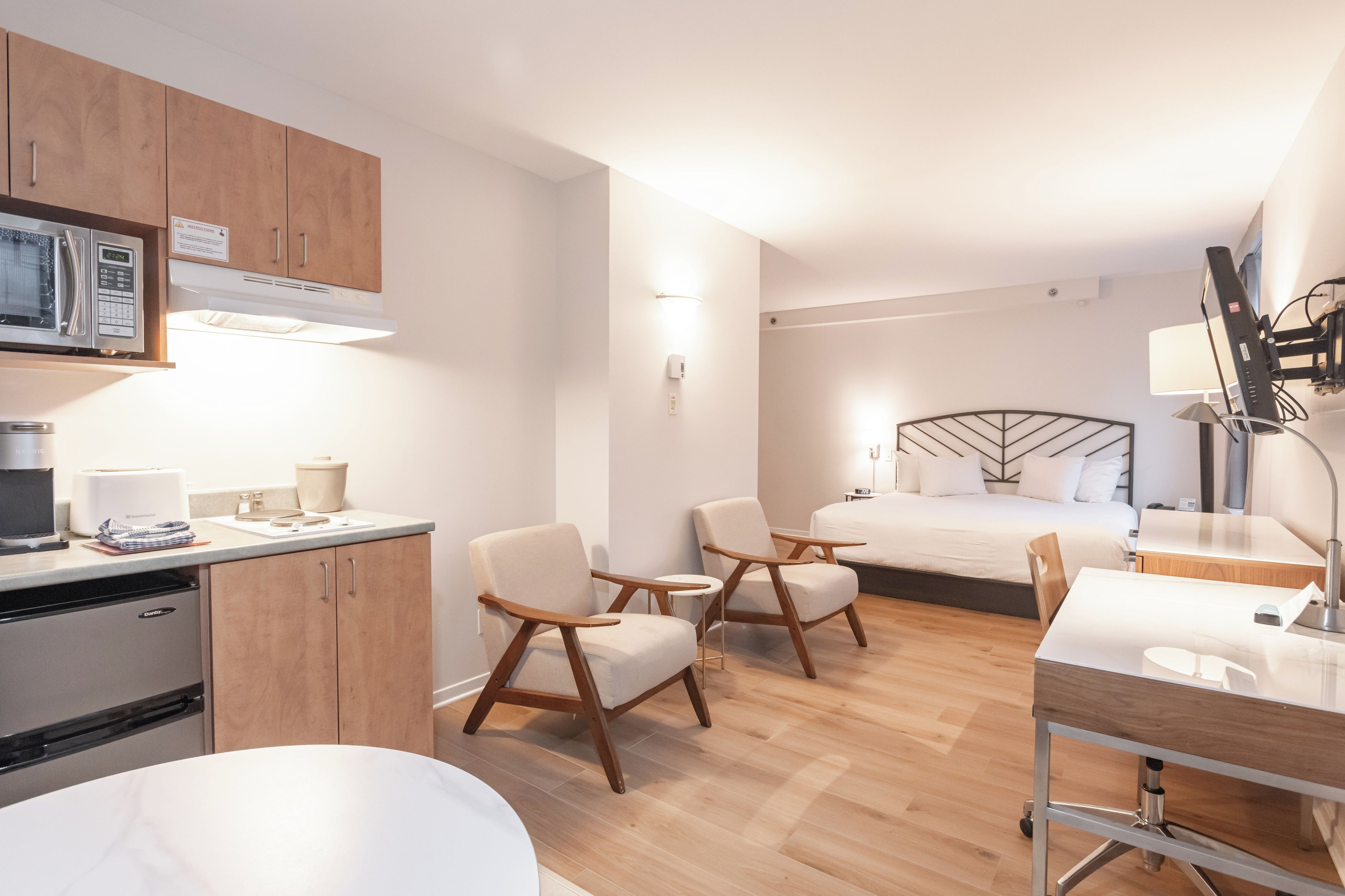 Ein modernes Hotelzimmer mit einer Küchenzeile, einem Bett, zwei Sesseln, einem Schreibtisch und einem Nachttisch.