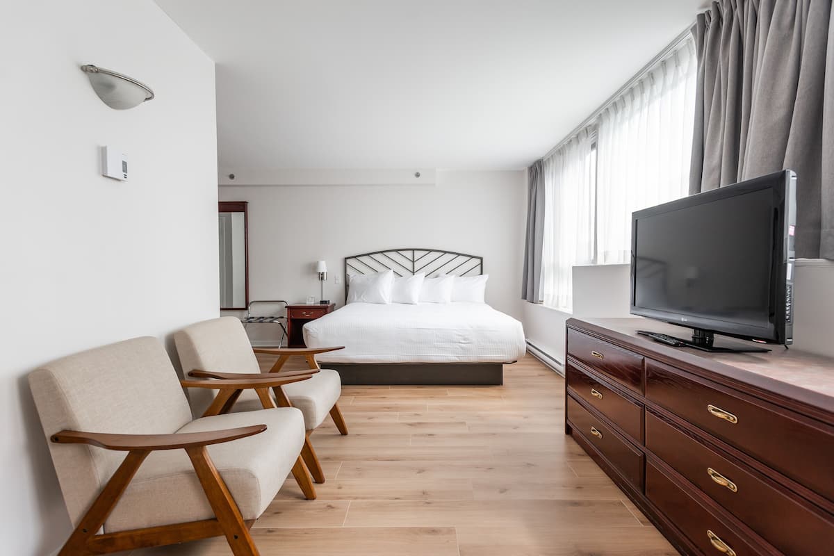 A modern hotel room with a bed, two armchairs, a TV, and a wooden dresser.