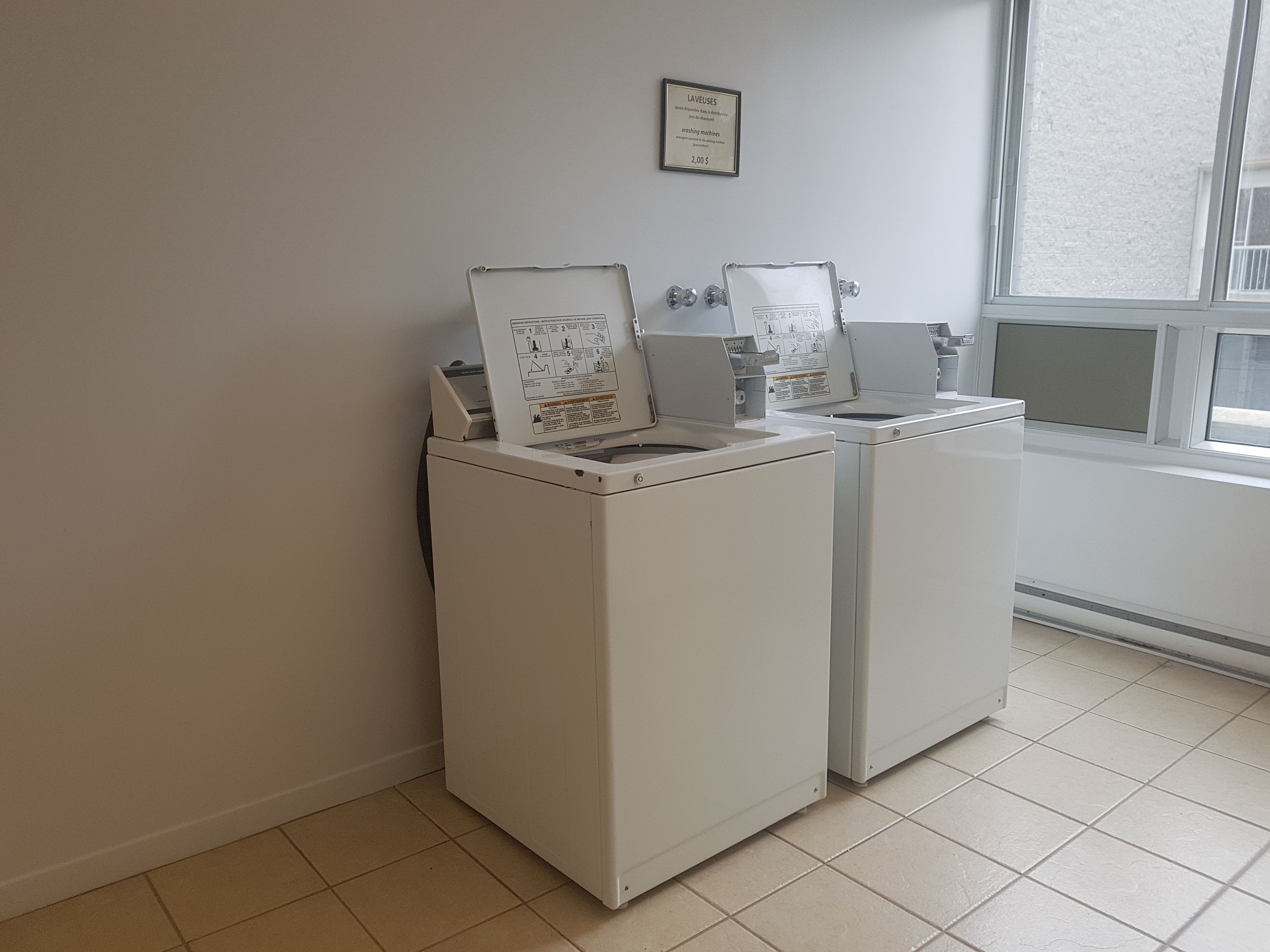 Een wasserij met twee witte wasmachines, een lijst met een bord aan de muur, en een raam dat natuurlijk licht binnenlaat.