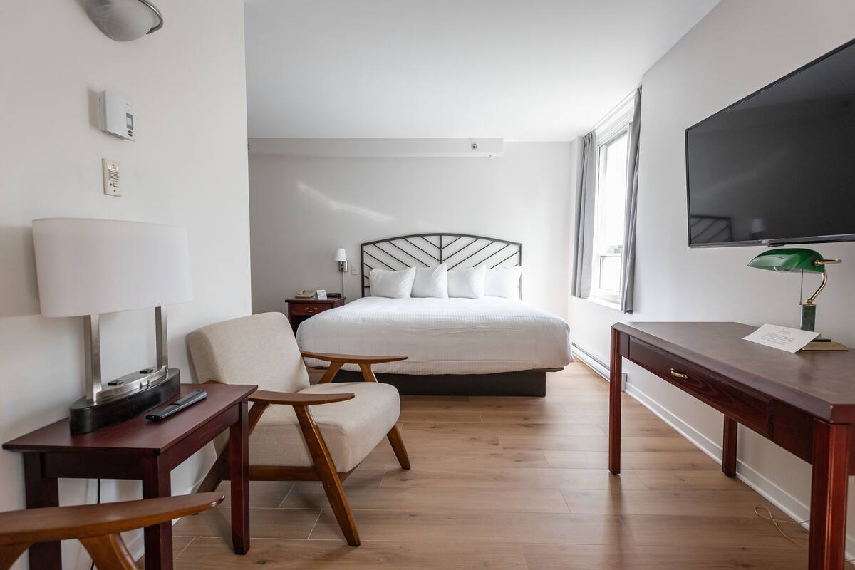 A hotel room with a bed, a desk, a chair, a TV, and a lamp.