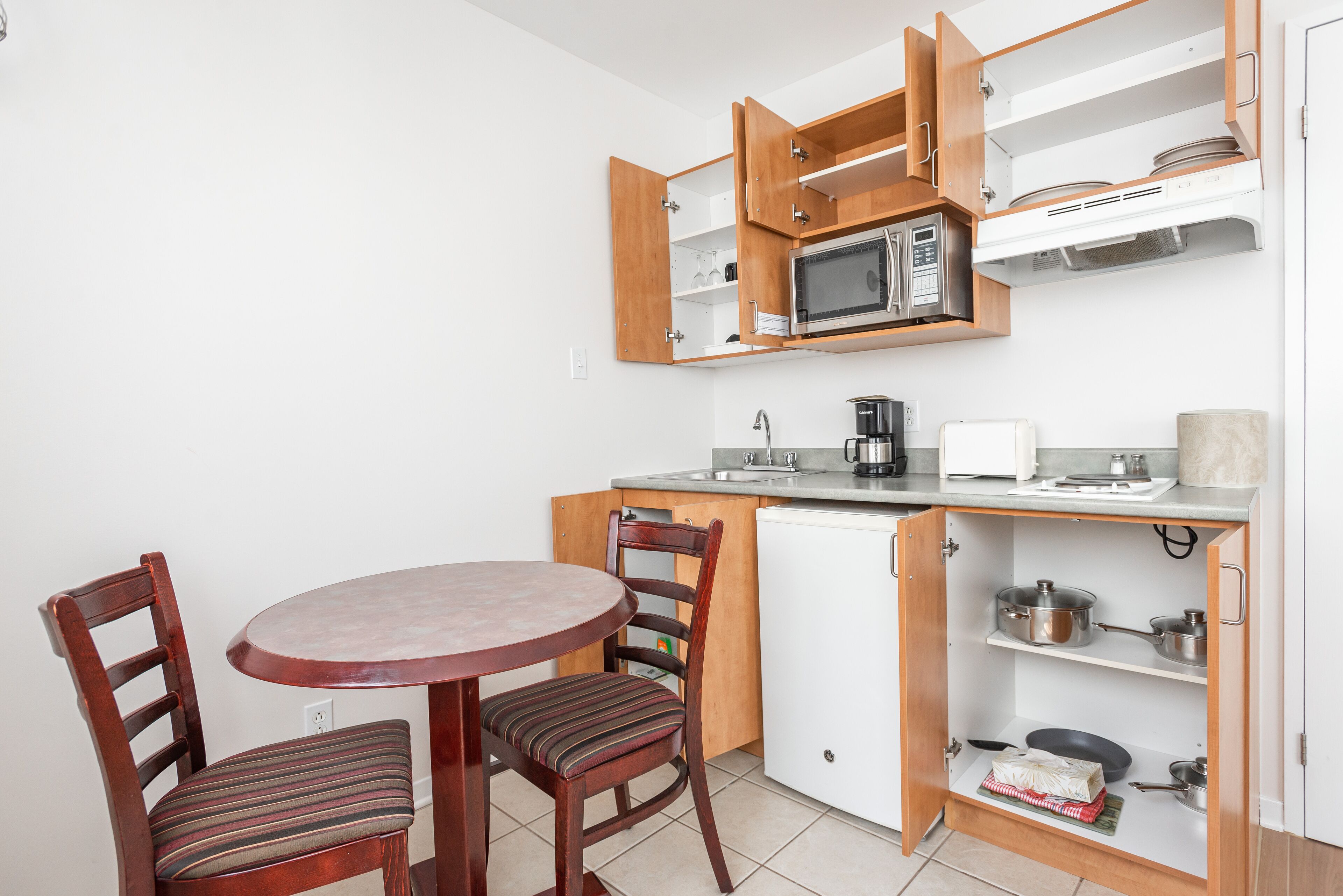 A compact kitchen with a microwave, coffee maker, and a small refrigerator. There is a dining table with two chairs and a cabinet with shelves above it.