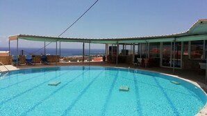 A swimming pool with clear blue water, surrounded by lounge chairs and a covered seating area with a view of the ocean. - Creta Suites (Ierapetra)
