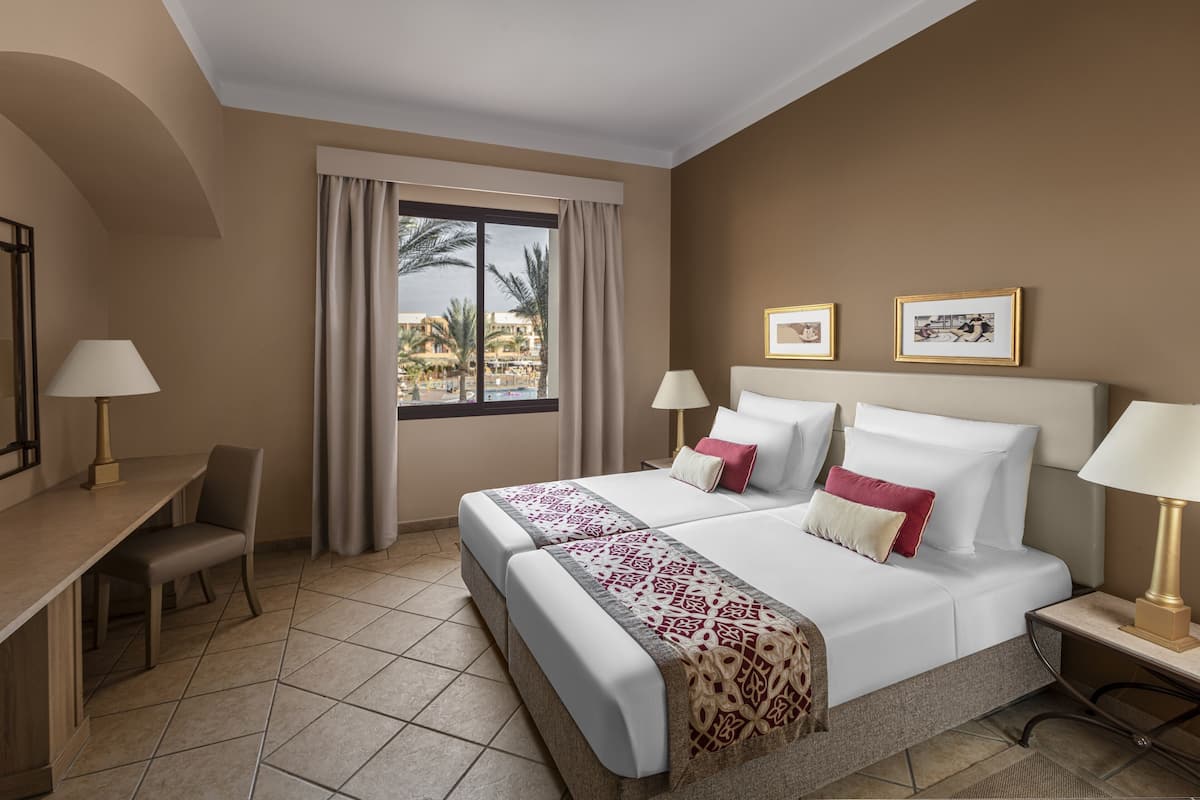 Una habitación de hotel con cama, dos mesitas de noche con lámparas, un escritorio con silla, una ventana con cortinas y cuadros en la pared.