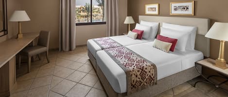 A hotel room with a bed, two bedside tables with lamps, a desk with a chair, a window with curtains, and framed pictures on the wall.
