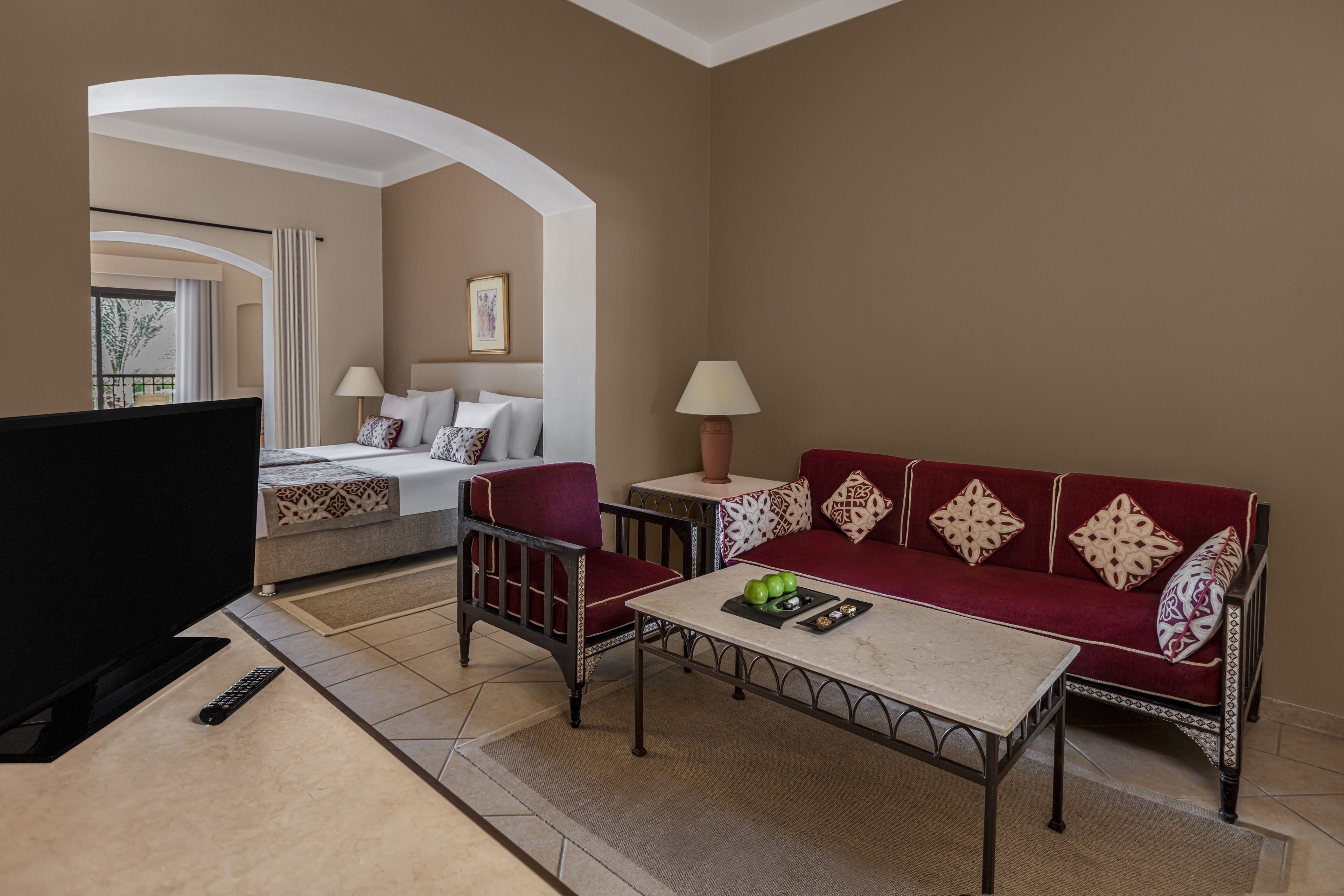 A hotel room with a TV, a sofa, a table with apples, and a bed with a lamp.