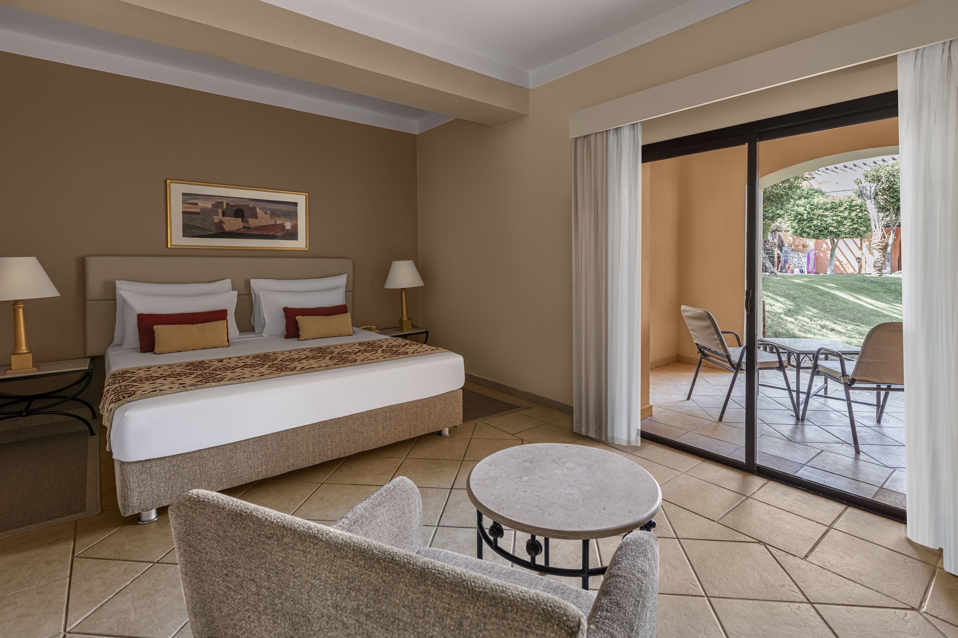 Habitación de hotel con cama, dos lámparas de noche, una mesa redonda y vistas a un patio exterior con sillas.