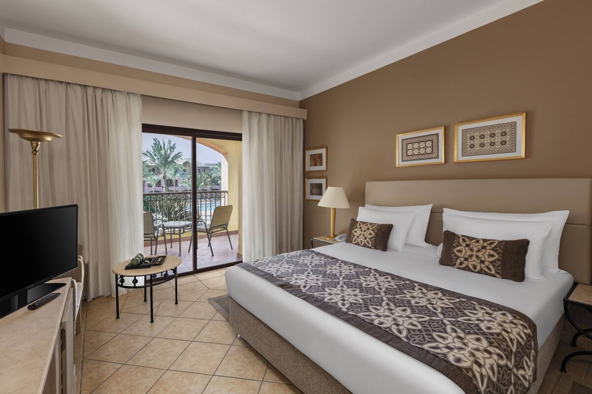 Habitación de hotel con una cama grande, televisión, escritorio y balcón con vistas a palmeras y edificios.
