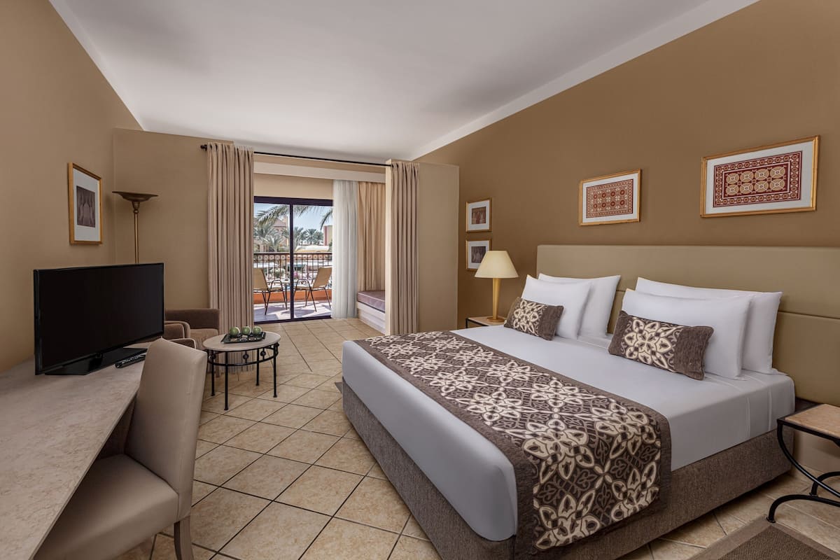Habitación de hotel con una cama grande, un escritorio con televisor, una mesita con lámpara y un balcón con cortinas.