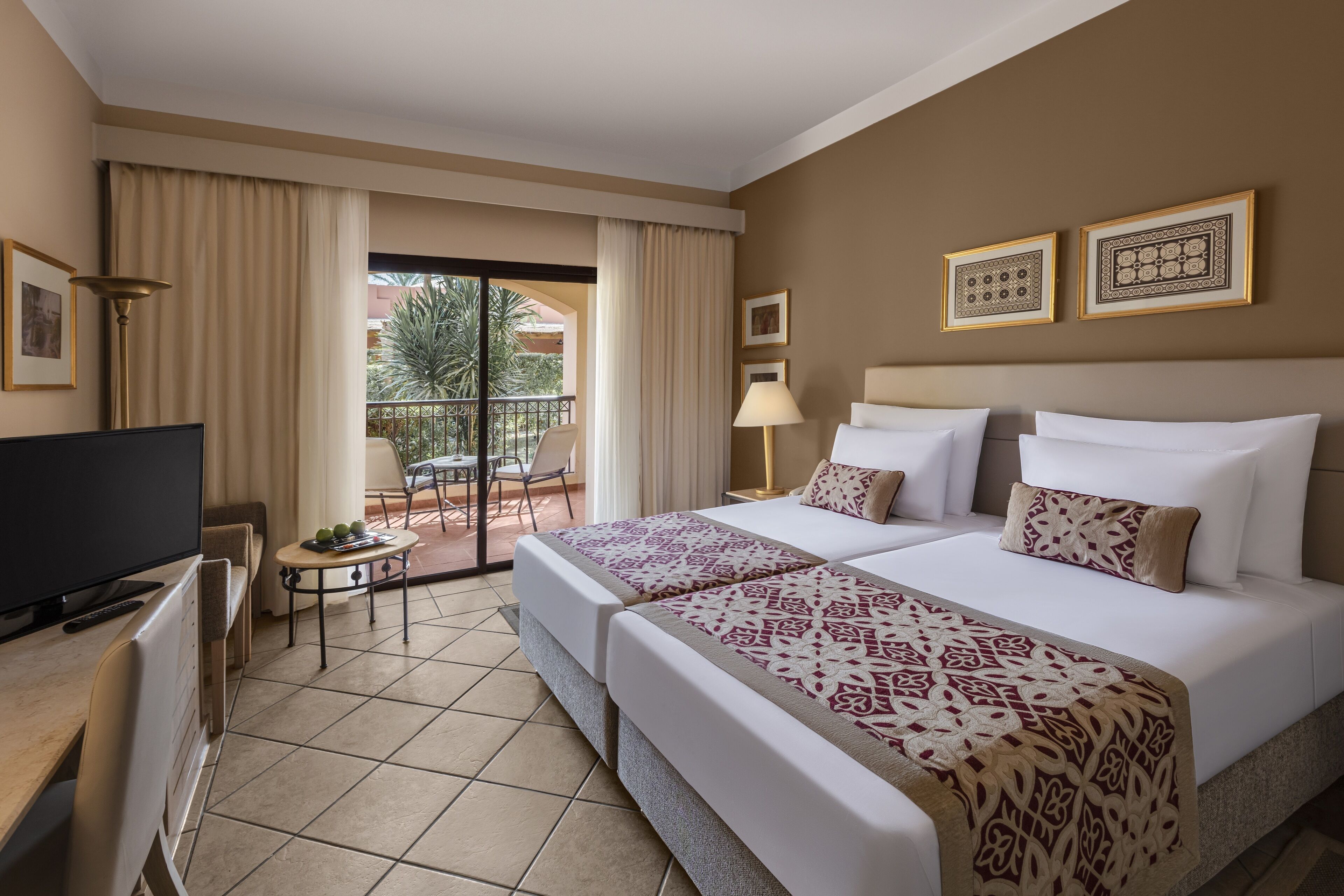 A hotel room with two beds, a TV, a small table with fruit, and a balcony with a view of palm trees.