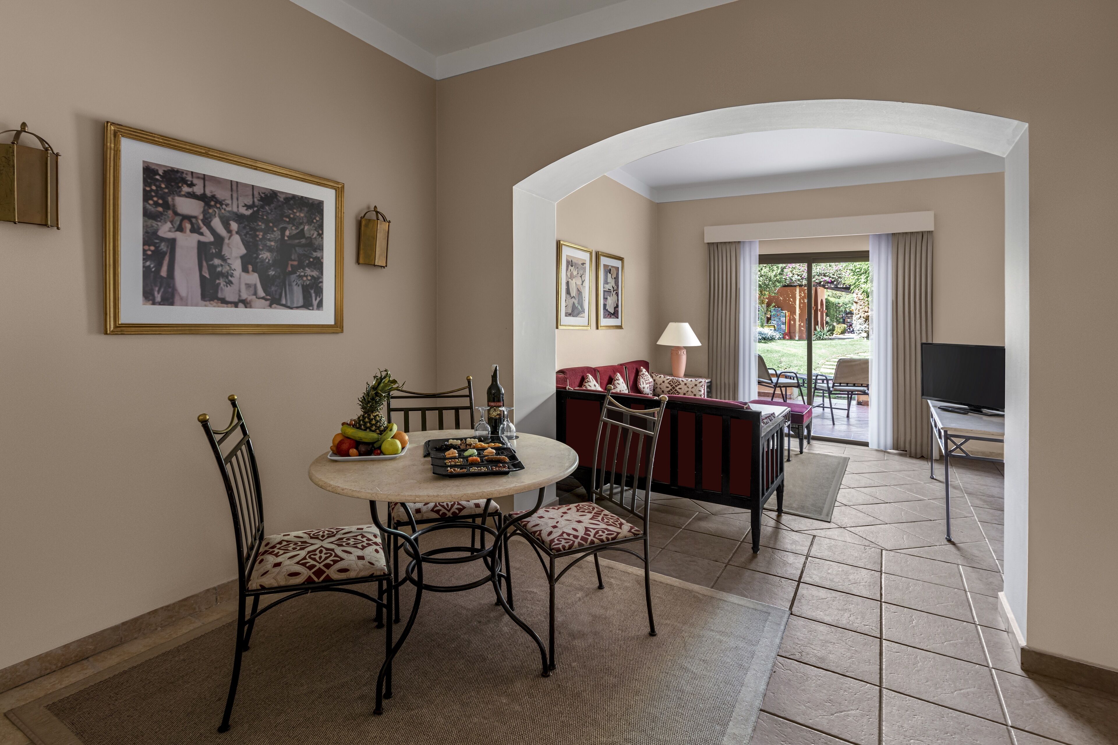 A dining area with a round table, chairs, and a small fruit basket. There is a television and a sofa in the background.