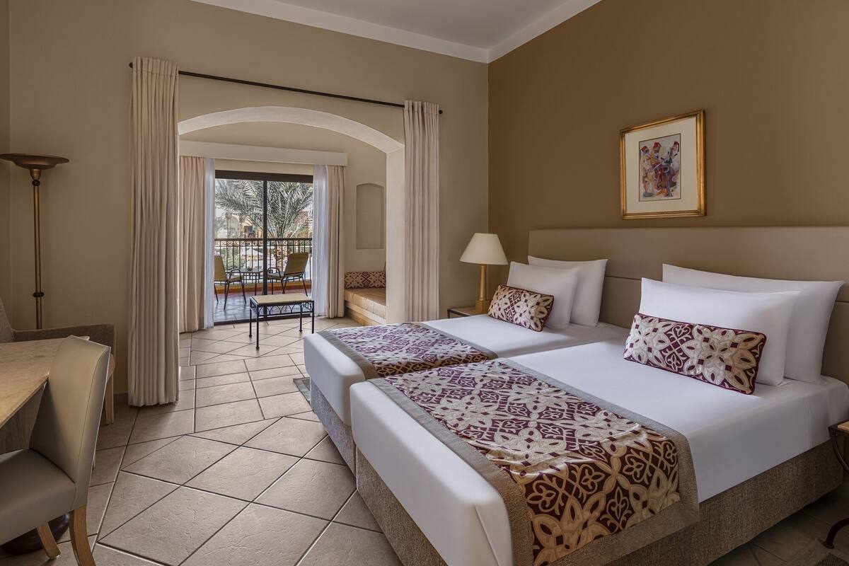 Habitación de hotel con dos camas, un balcón con sillas, un escritorio y una lámpara.