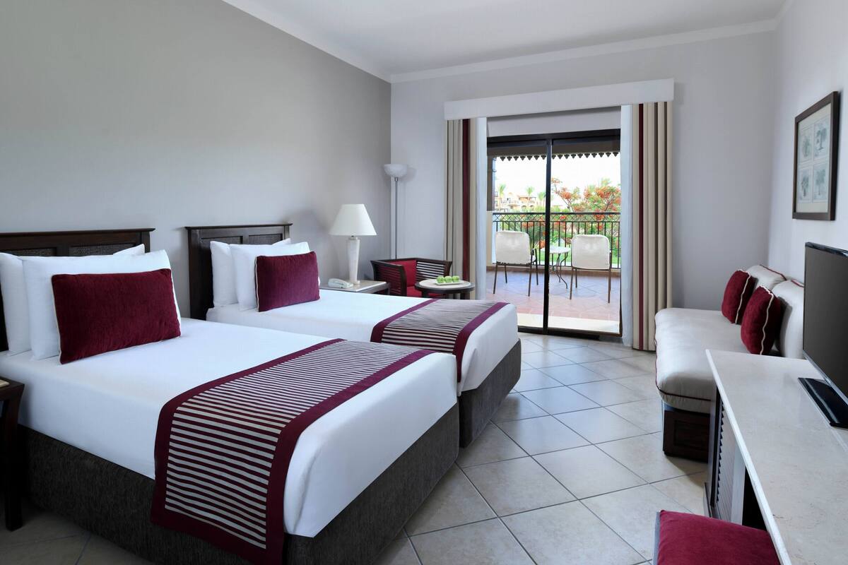 A hotel room with two beds, a desk, a chair, a small table, and a balcony with outdoor seating.