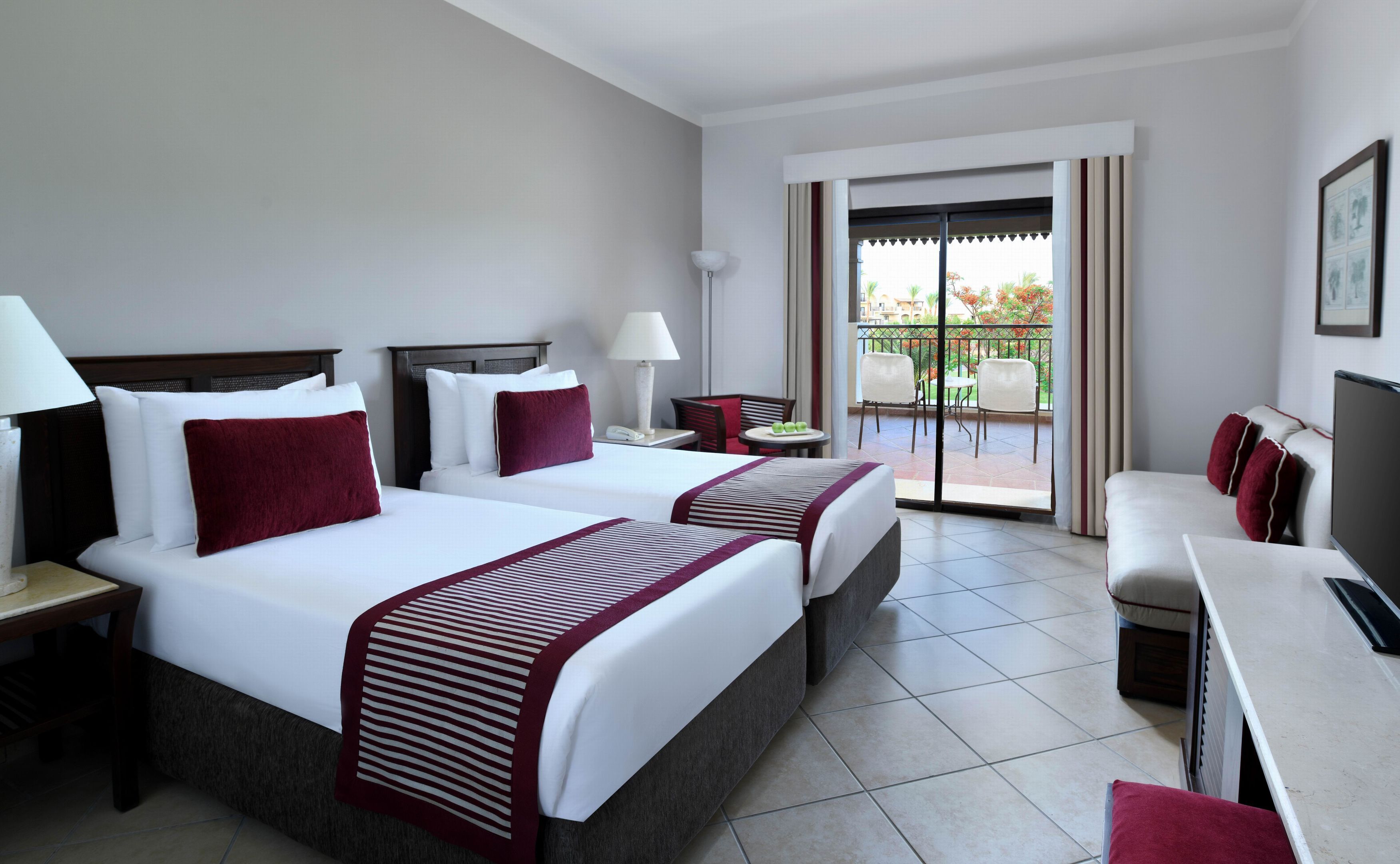 A hotel room with two beds, a desk, a chair, a small table, and a balcony with outdoor furniture.