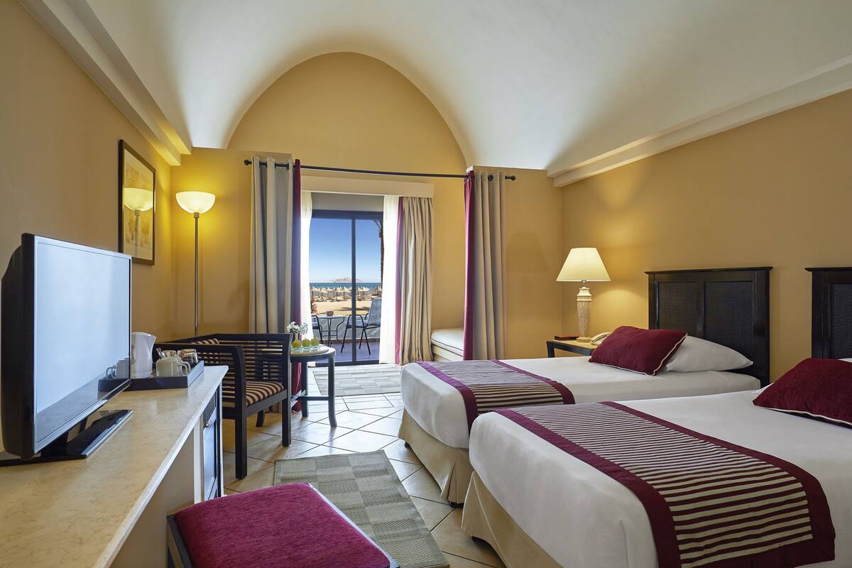 A hotel room with two beds, a desk with a TV, a chair, a lamp, and a view of the outdoors.