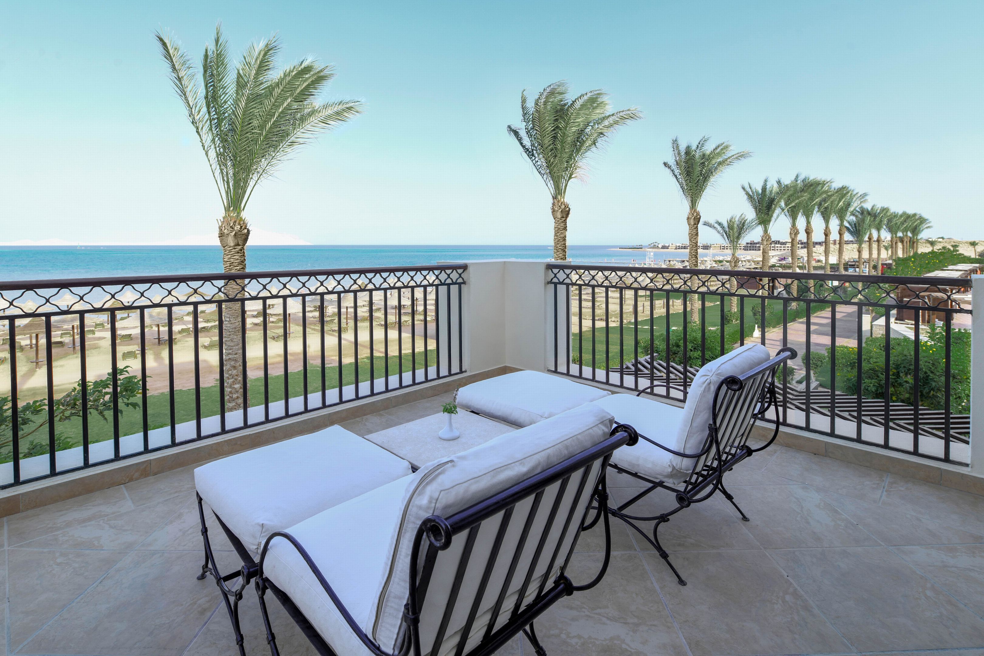 Un balcon avec deux chaises blanches dotées de coussins et une petite table, donnant sur une plage bordée de palmiers et surplombant un ciel dégagé.