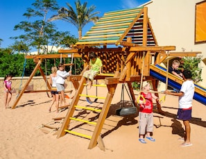 Une structure de jeux en bois, dotée de toboggans et d’éléments d’escalade, installée sur une surface sablonneuse.