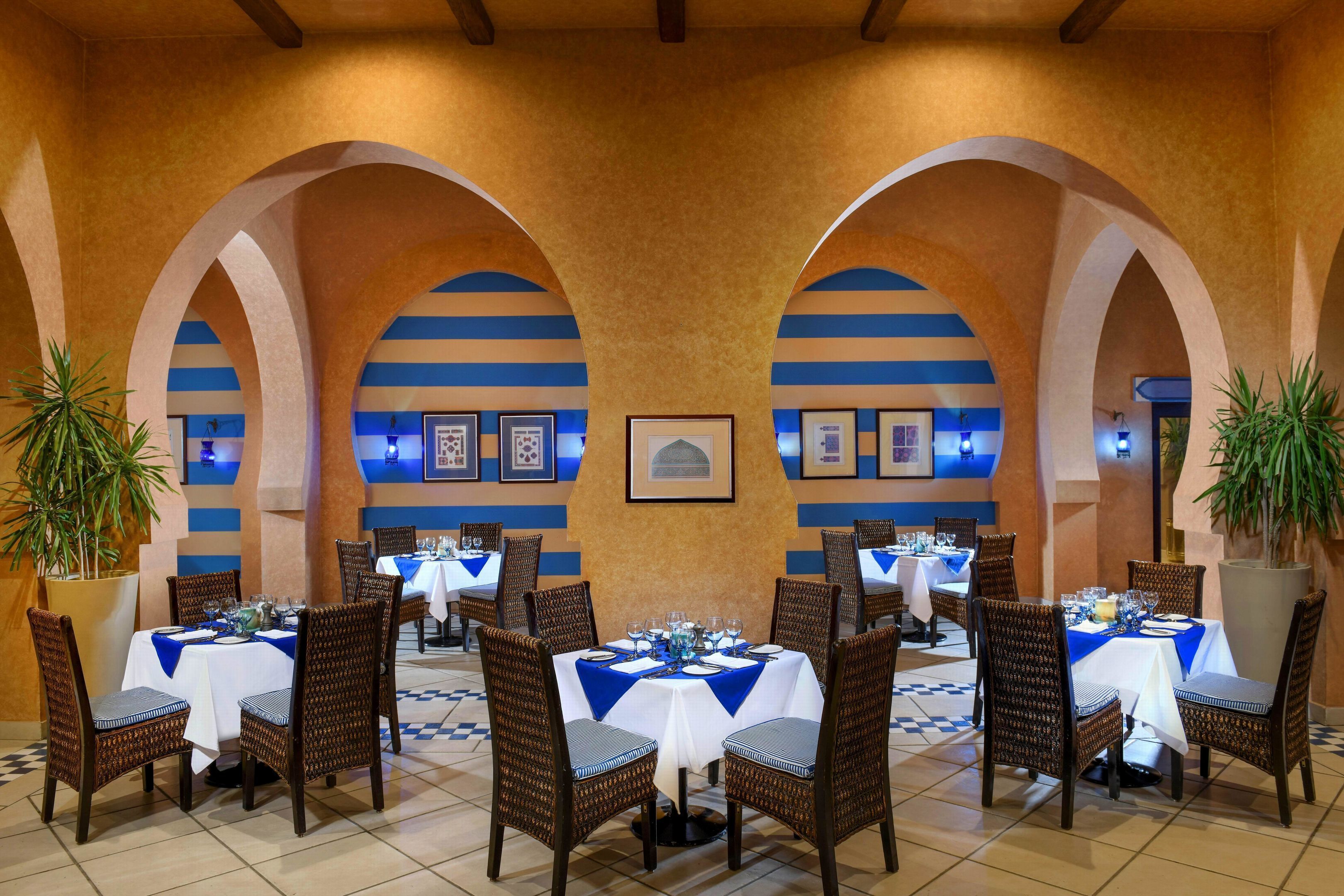Zona pranzo con tavoli apparecchiati per quattro, ognuno con tovaglia bianca e dettagli blu, circondati da sedie in vimini.