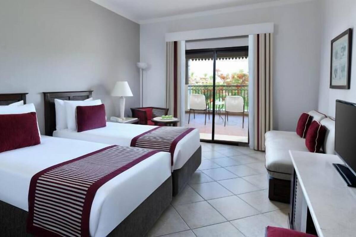 A hotel room with two beds, a desk, a TV, and a balcony with outdoor seating.