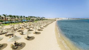 Una playa con hileras de tumbonas y sombrillas, una piscina y un edificio de resort al fondo.