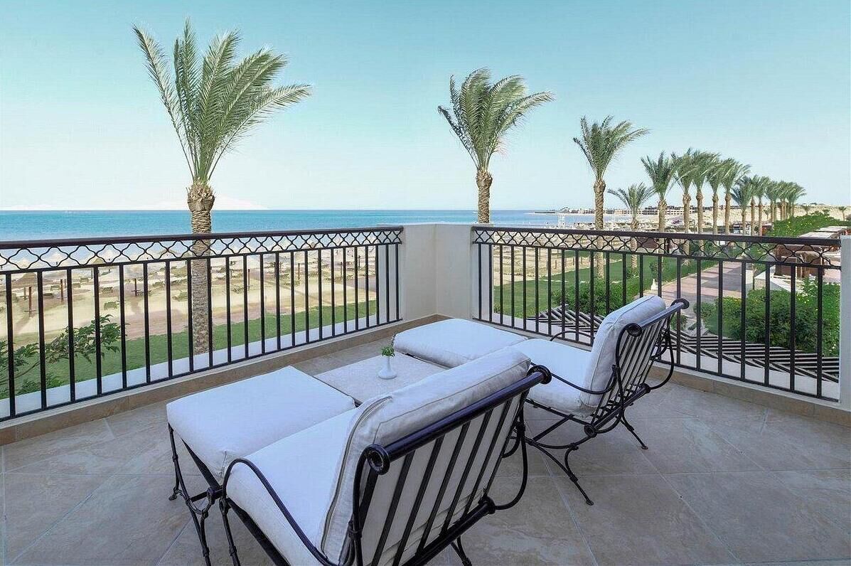 A balcony with two white cushioned chairs and a small table, overlooking a beach with palm trees and a clear sky.
