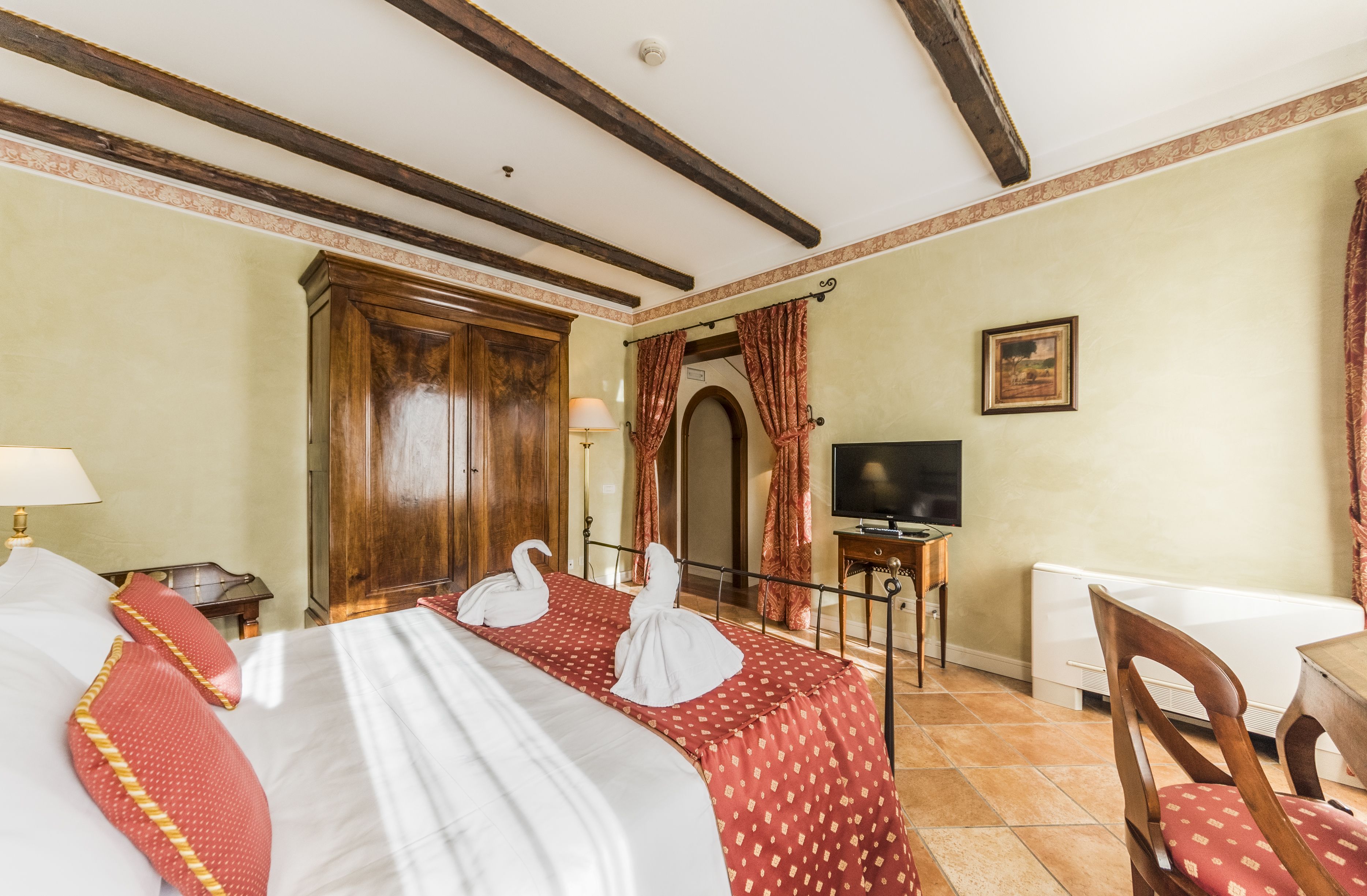 A hotel room with a large bed, two red pillows, a wooden wardrobe, a television, and a desk with a chair.