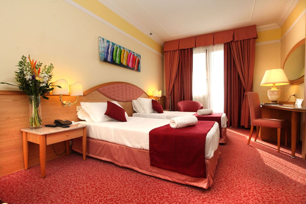 Une chambre d’hôtel avec deux lits, un bureau, une chaise, une lampe, un tableau et un vase avec des fleurs.