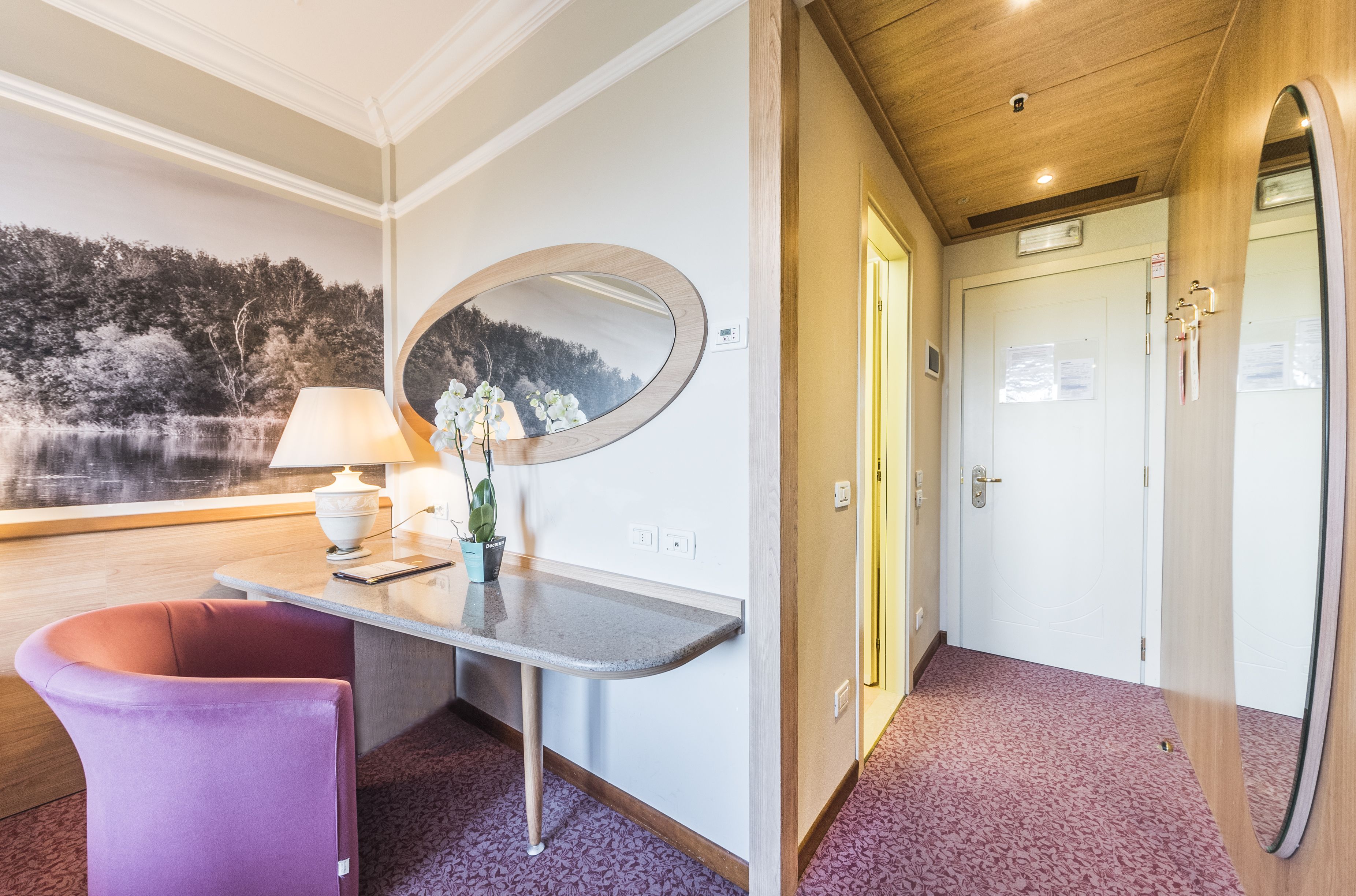 A room with a wooden desk, a purple chair, a mirror, and a view of a lake.
