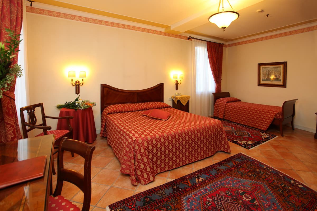 Une chambre d’hôtel avec un grand lit, un plus petit lit, une table à manger et des chaises. Il y a deux lampes fixées au mur et un tableau accroché au mur.