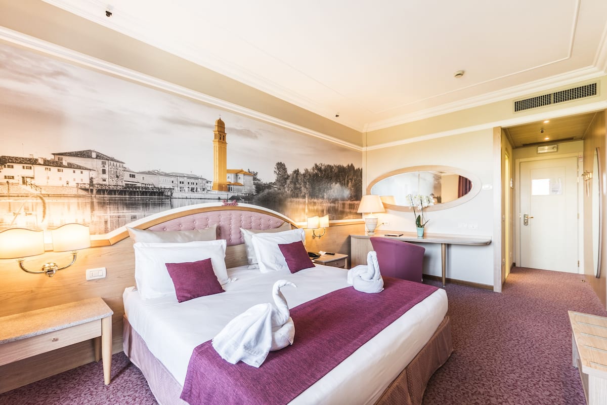Une chambre d’hôtel équipée d’un lit, de tables de chevet, d’un bureau et d’une grande fenêtre donnant sur le paysage urbain.