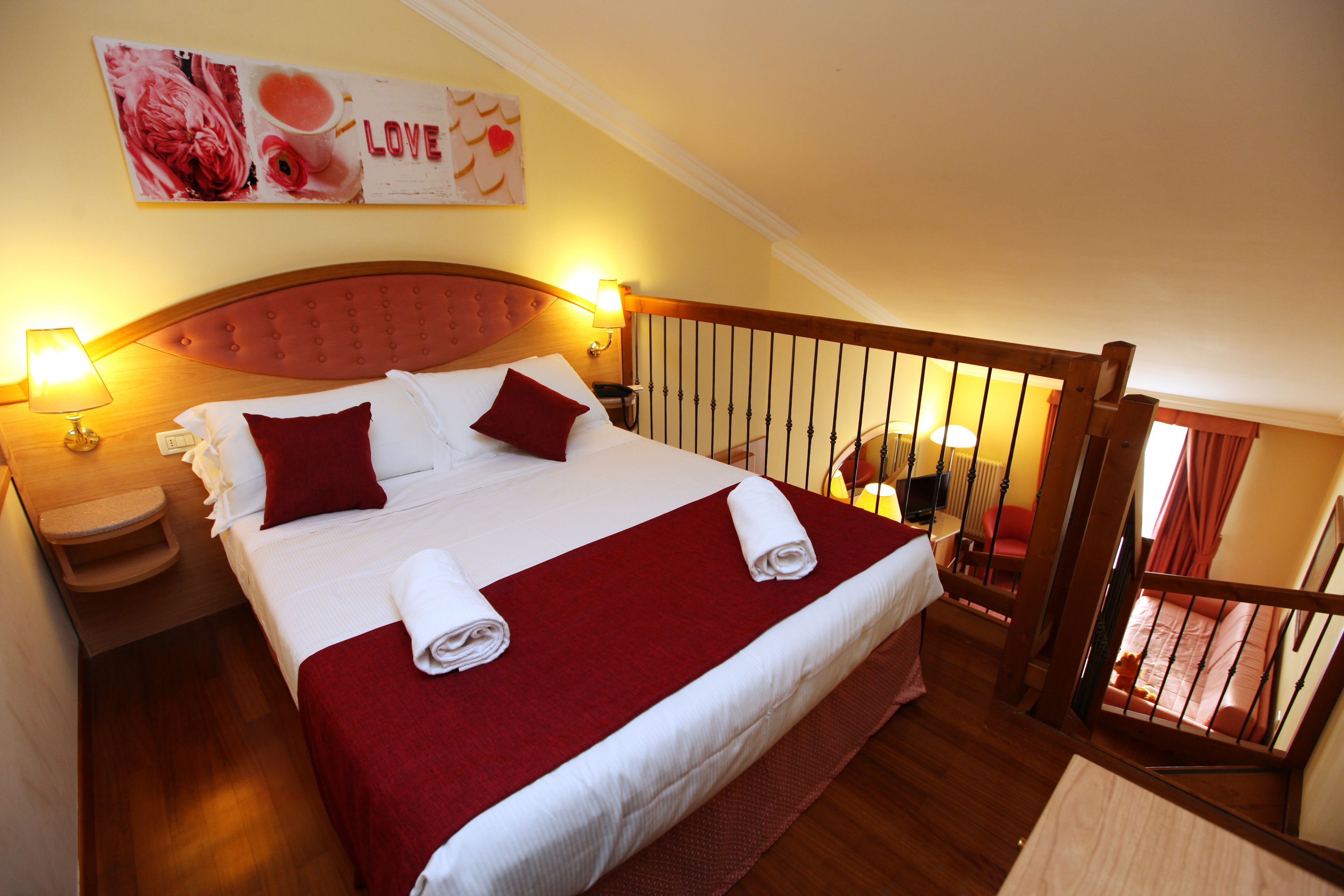 Habitación de hotel con una cama grande, ropa de cama roja, cabecera de madera, dos lámparas de noche, una mesita y vistas a un balcón con barandilla.