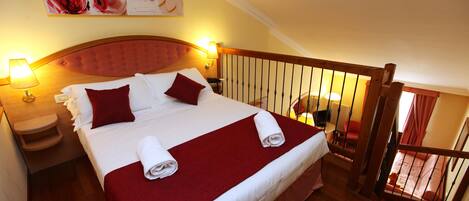 A hotel room with a large bed, red bedding, a wooden headboard, two bedside lamps, a small table, and a view of a balcony with a railing.