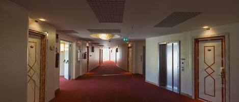 A hotel corridor with a red carpet, multiple doors, and a ceiling with recessed lighting.