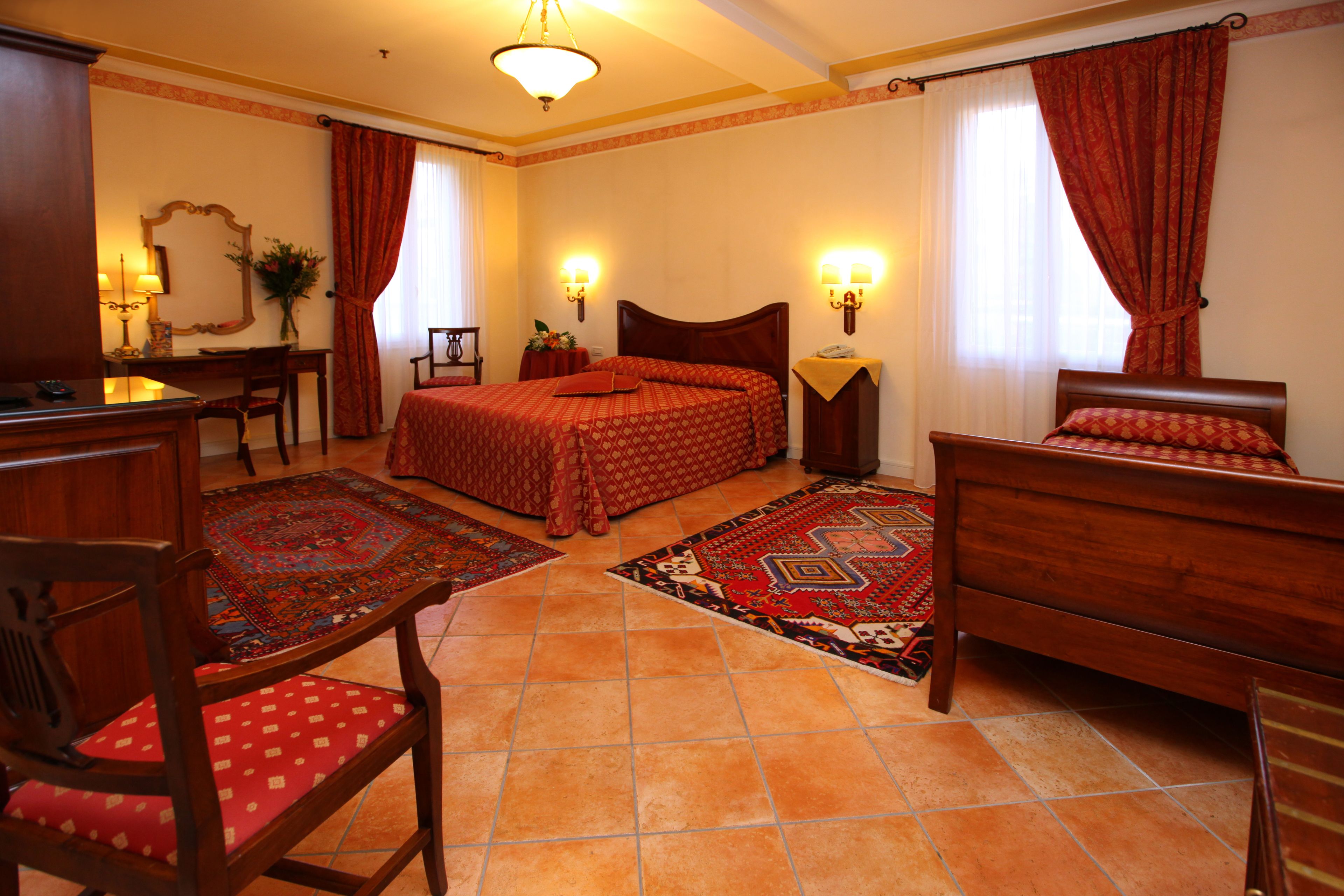 Una habitación de hotel con una cama, dos sillas de madera, un escritorio con una lámpara, un espejo y dos lámparas de pared.