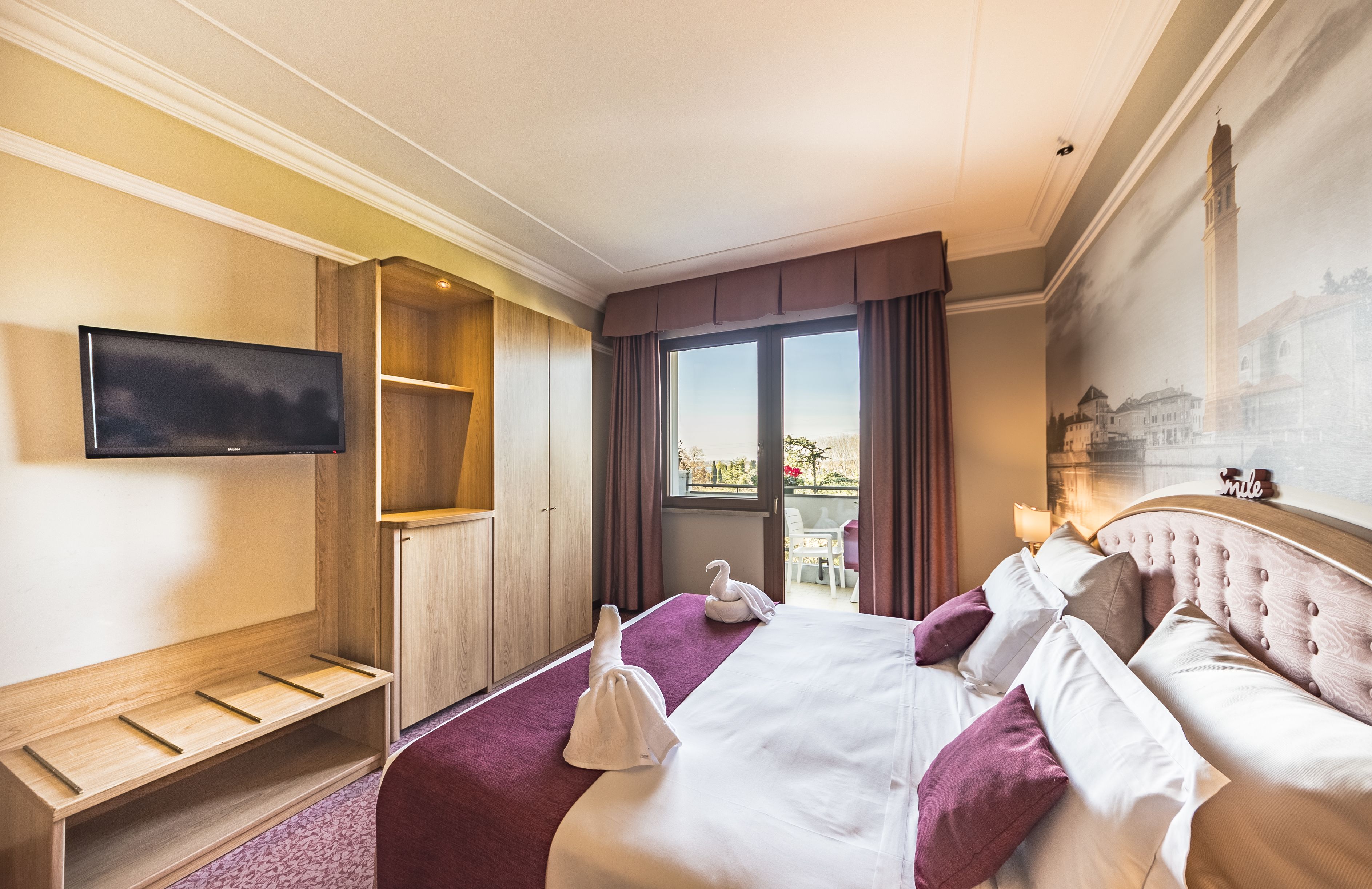 A hotel room with a large bed, a television, a wooden bench, and a view of the city.