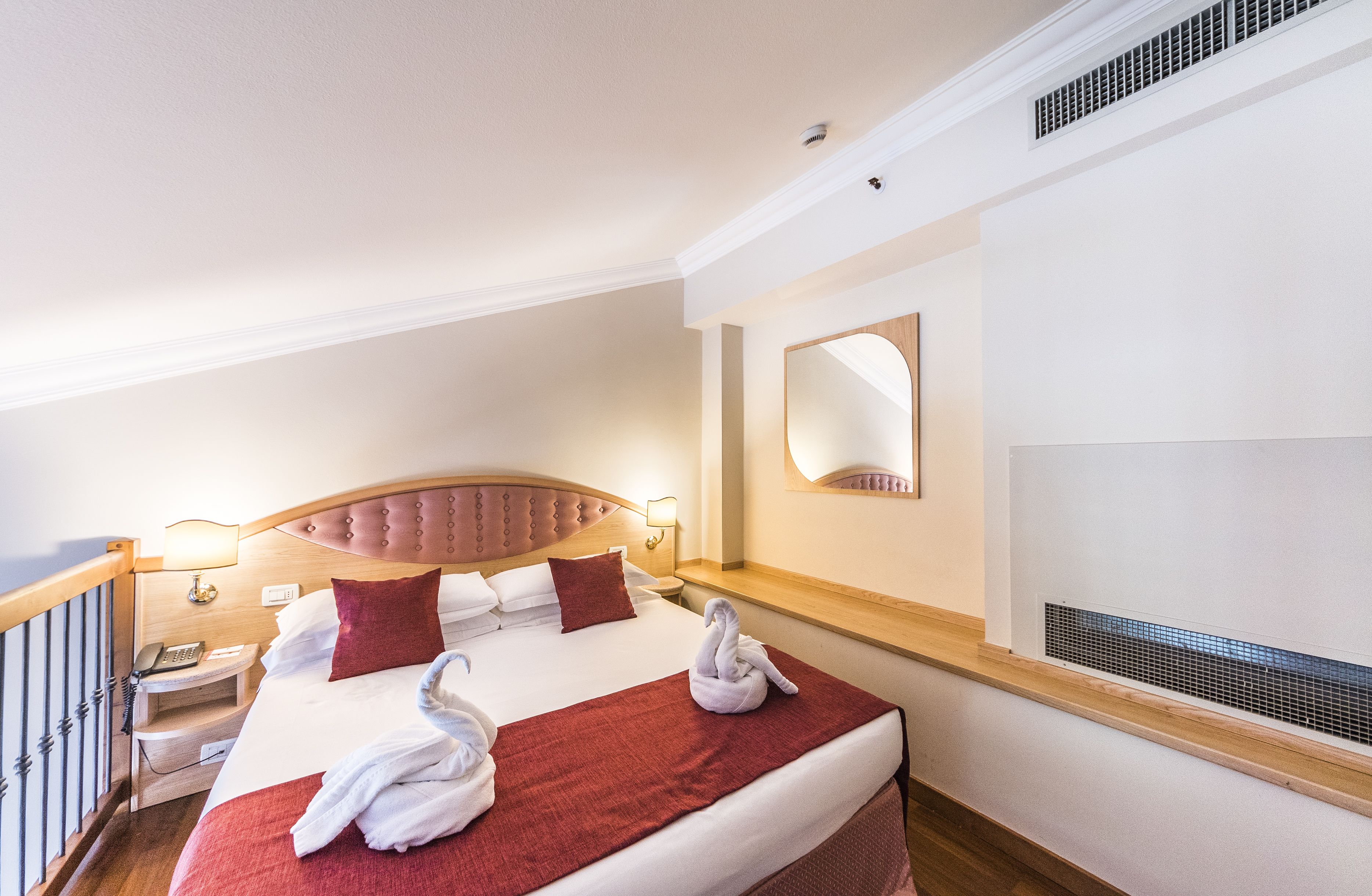 Habitación de hotel con una cama grande, un cabecero de madera, una mesita de noche con lámpara, un espejo y un respiradero en la pared.