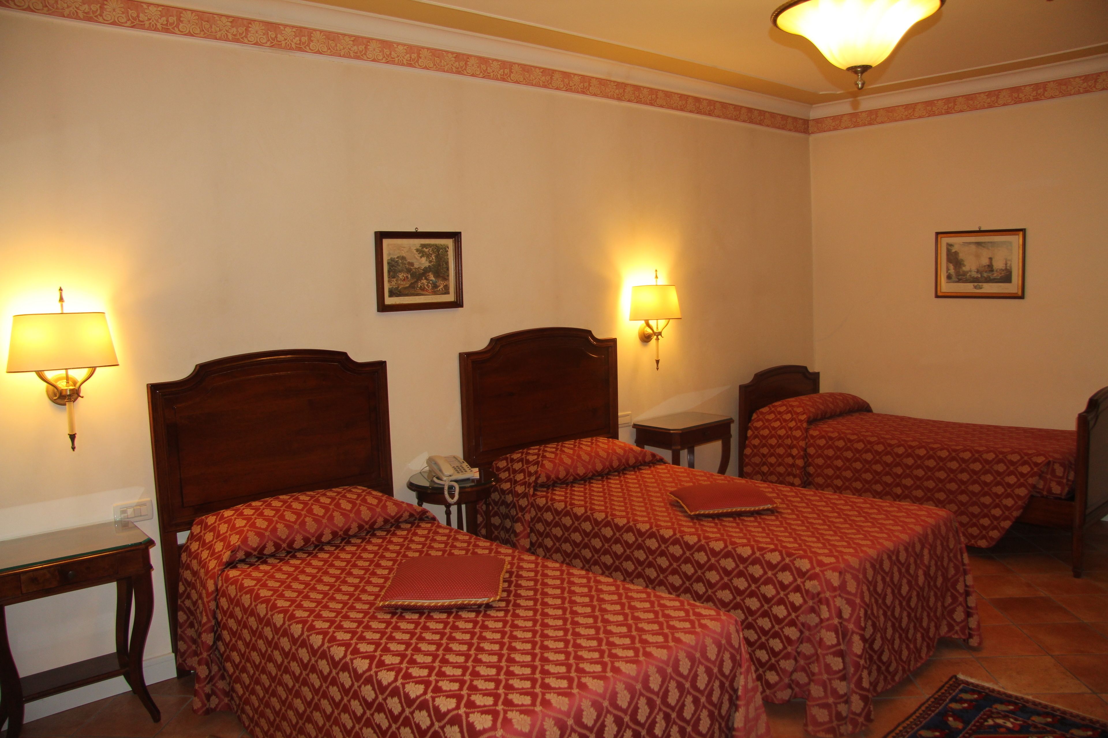 A hotel room with three beds, each with a patterned bedspread, a bedside table with a lamp, and a small table with a telephone.
