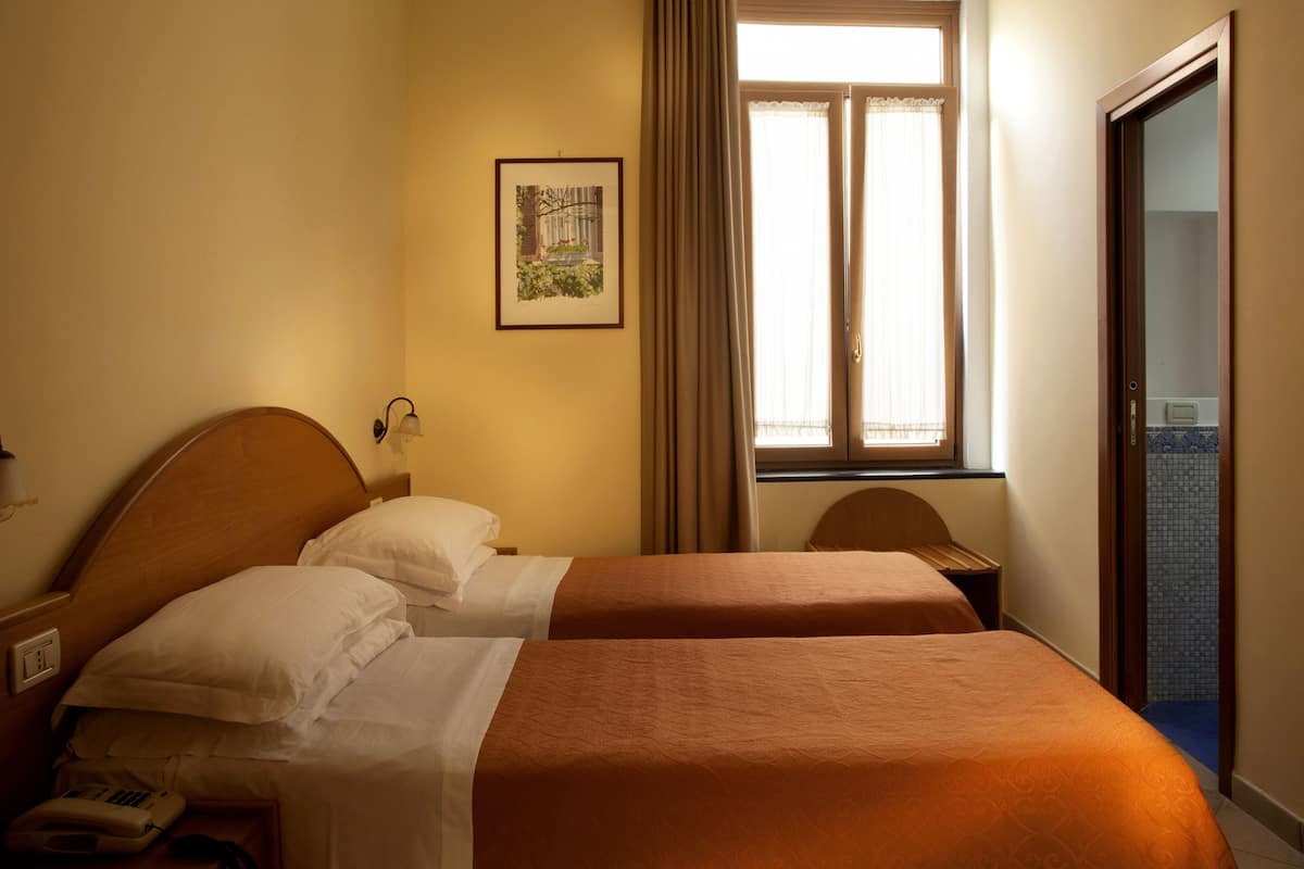A hotel room with two beds, a window with curtains, a wall-mounted light, and a framed picture.