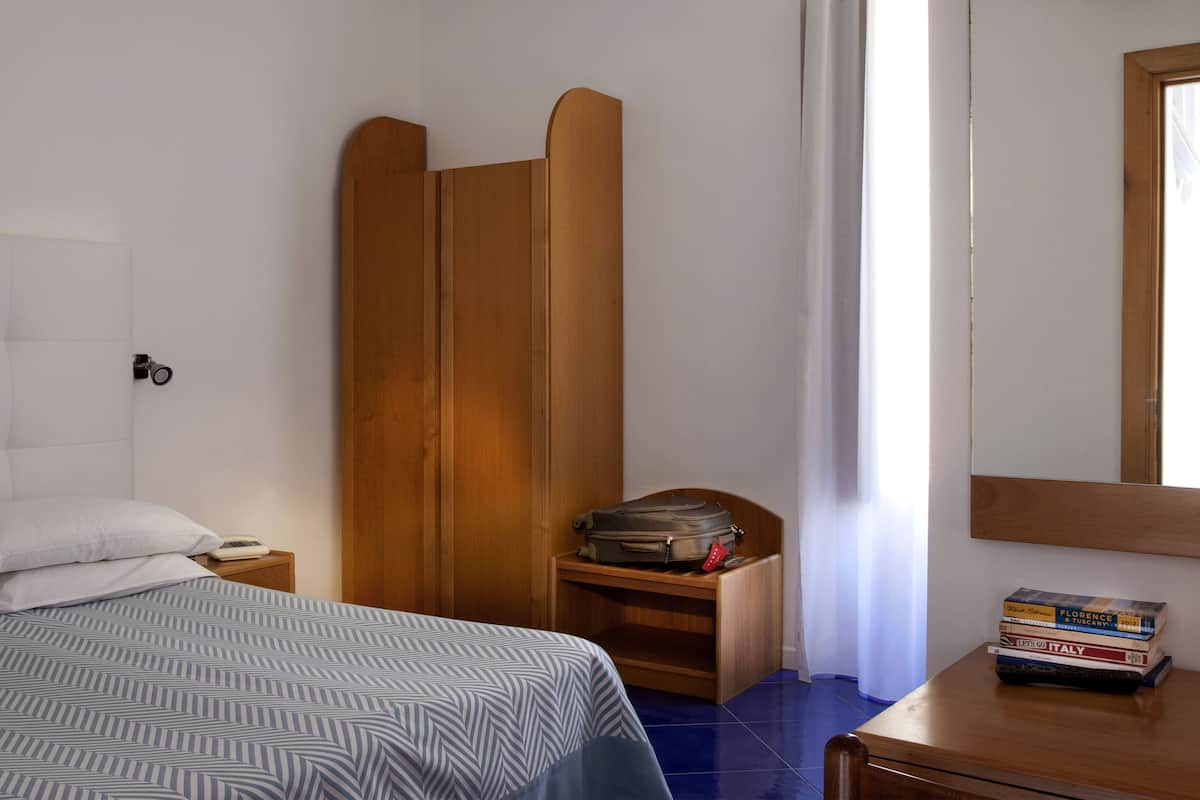 A hotel room with a bed, a wooden wardrobe, a bedside table with a lamp, and a stack of books.