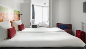 A hotel room with a large bed, a desk, a chair, a TV, and a red suitcase.