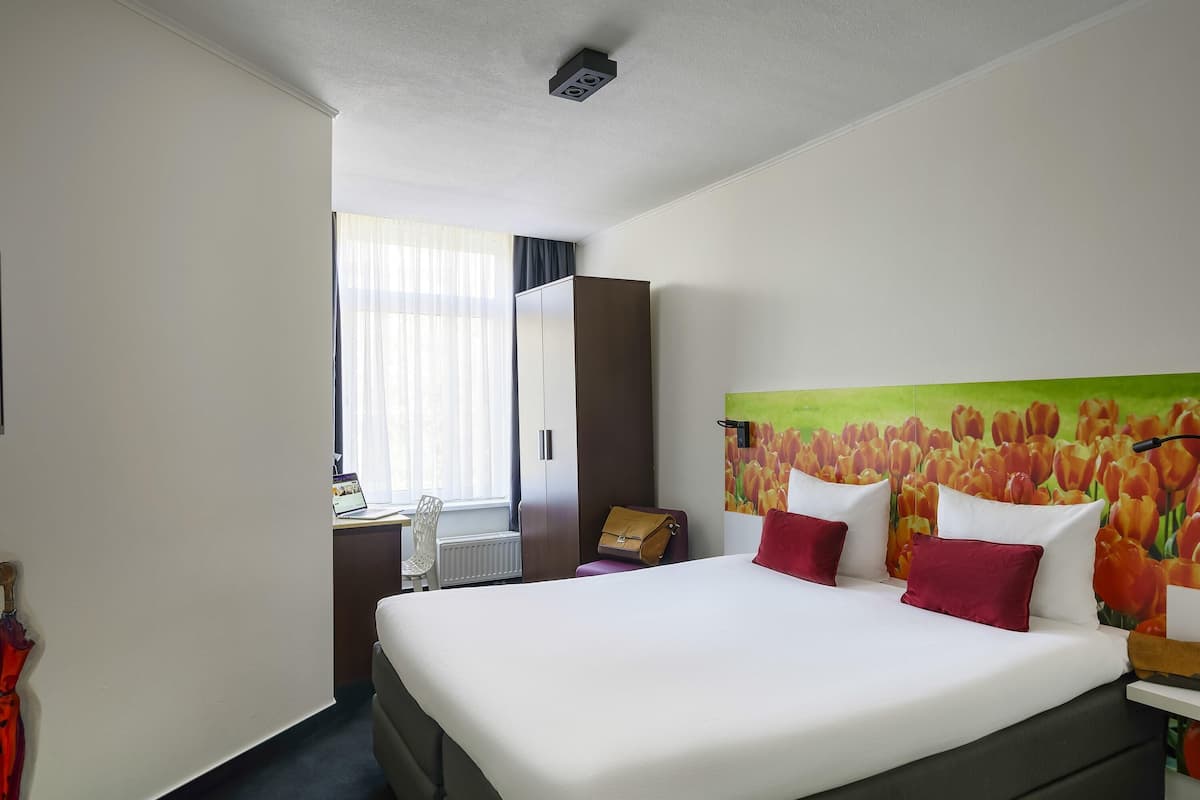 A hotel room with a bed, a desk, a chair, a radiator, and a painting of tulips on the wall.