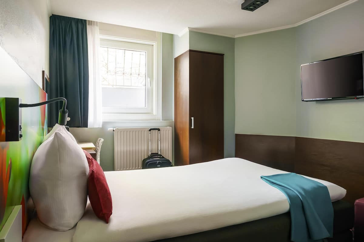 A hotel room with a single bed, a TV mounted on the wall, a radiator, and a window with curtains.