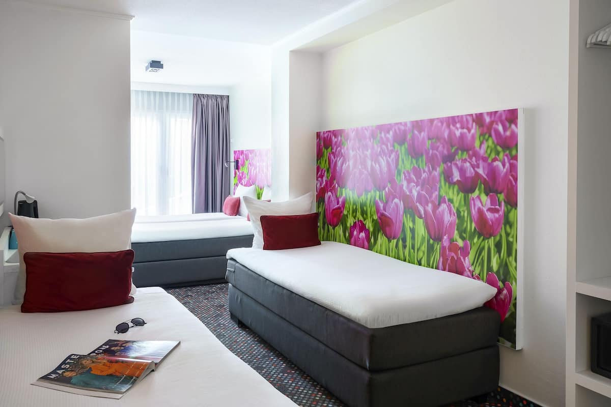 A hotel room with a bed, a sofa, a large painting of flowers, and a window with curtains.