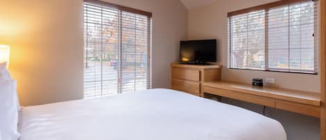A hotel room with a large bed, a TV on a wooden shelf, and two windows with blinds.