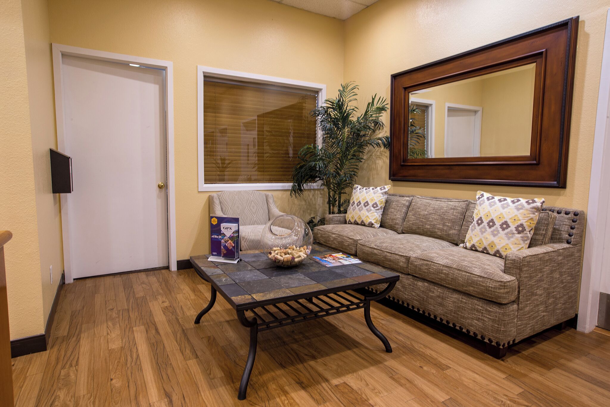A living room with a wooden floor, a sofa, a coffee table, a window with blinds, a door, and a large mirror.